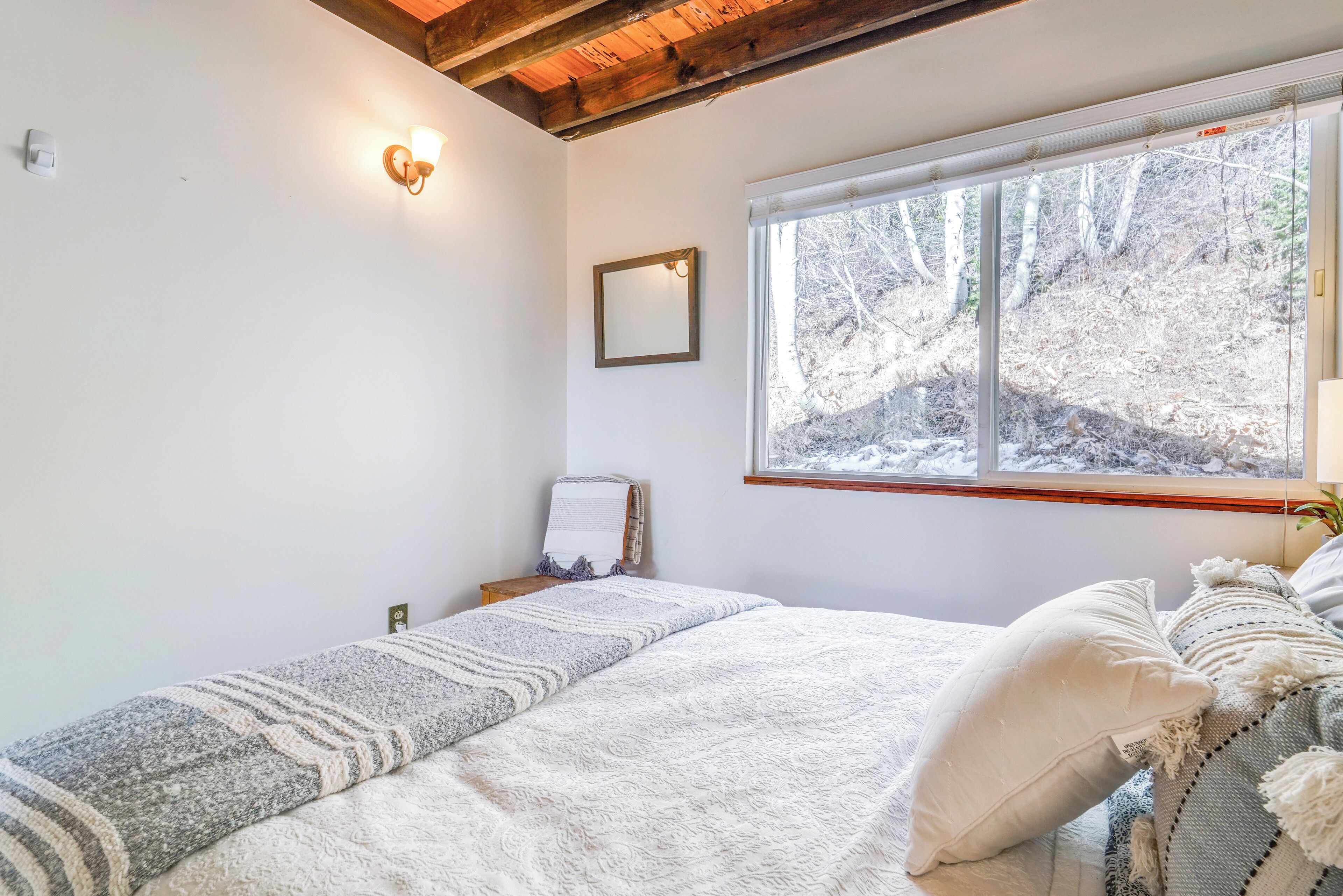 Une chambre à coucher avec un lit, une fenêtre donnant sur des arbres, une lampe fixée au mur et une petite table de chevet.