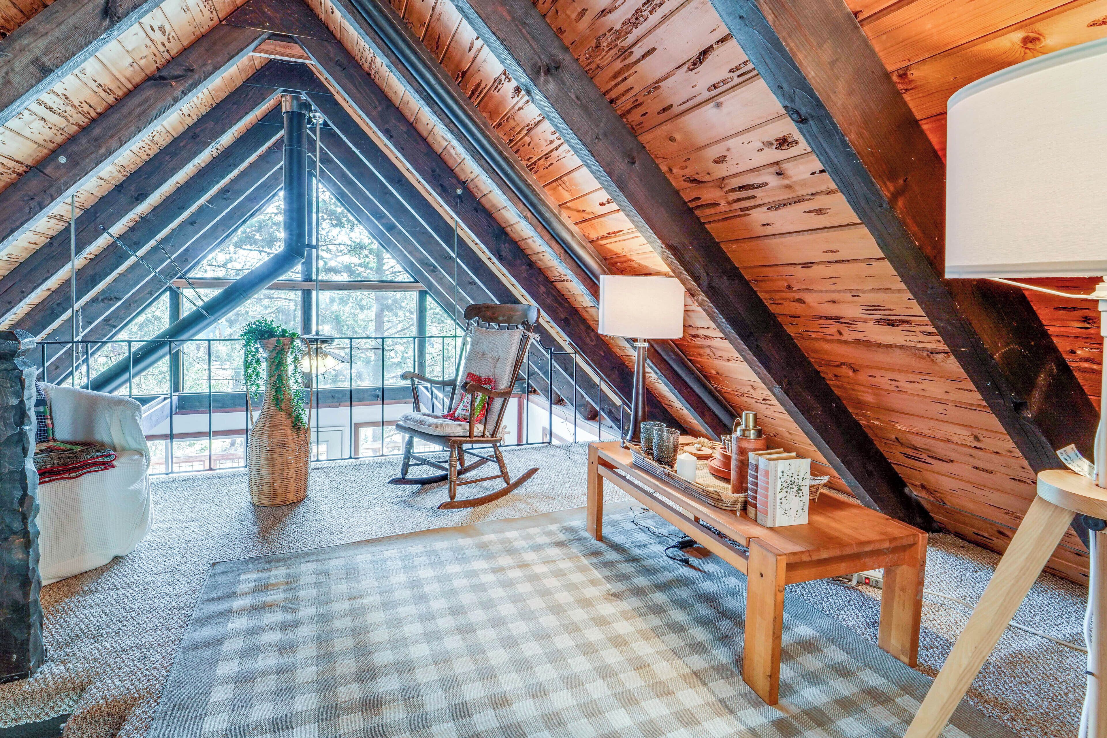 An attic room with a high ceiling, wooden beams, a rocking chair, a table with books, and a window with a view of trees.
