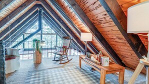 Una habitación en el ático con techo alto, vigas de madera, una mecedora, una mesa con libros y una ventana con vista a árboles.