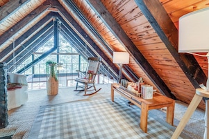 Una habitación en el ático con techo alto, vigas de madera, una mecedora, una mesa con libros y una ventana con vista a árboles.
