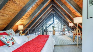 Un dormitorio acogedor en el ático, con techo alto, cama grande con ropa de cama roja y blanca, sillón mecedor y vistas al exterior.