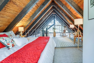 Un dormitorio acogedor en el ático, con techo alto, cama grande con ropa de cama roja y blanca, sillón mecedor y vistas al exterior.