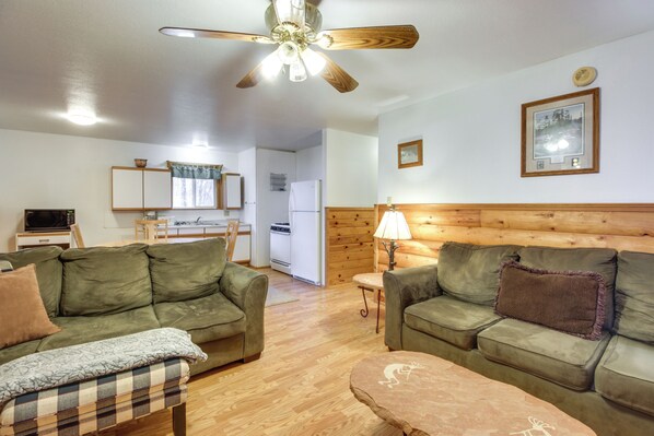 A cozy living room with two green sofas, a coffee table, and a ceiling fan. A kitchen with a refrigerator and microwave is visible in the background.