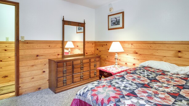 A room with a wooden headboard bed, a wooden dresser with drawers, a mirror, and a bedside table with a lamp. - Lakefront Bemidji Getaway w/ Community Beach! (Bemidji)