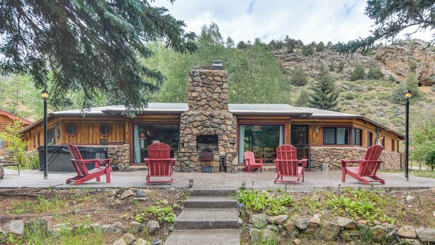 Une cheminée en pierre, entourée de fauteuils Adirondack rouges, se trouve devant une cabane en bois dotée d’une cheminée en pierre.