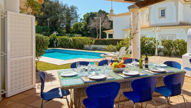 An outdoor dining area with a pool view, blue chairs, and a table set for four. - Villa Costa Rica - Four Bedroom Villa, Sleeps 8 (Algarve)