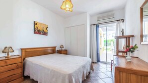A bedroom with a wooden bed, a nightstand, a dresser, a lamp, a painting, and a view of the outside through a glass door. - Villa Ninos - Three Bedroom Villa, Sleeps 6 (Albufeira)