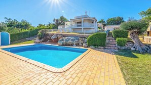 En swimmingpool med en terrass av tegelsten, omgiven av en vackert anlagd trädgård med stenar och buskar, och ett stort hus med en balkong i bakgrunden.