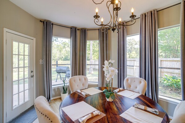 Uma sala de jantar com uma mesa redonda, quatro cadeiras, um candelabro e janelas amplas com cortinas.