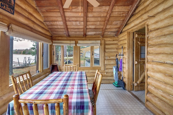 Interior de una cabaña con una mesa grande, sillas de madera, un ventilador de techo y vistas al exterior.
