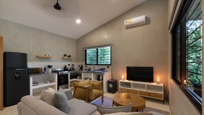 A modern living room with a sofa, a coffee table, and a TV. There is a kitchen area with a refrigerator, stove, and microwave. A window provides a view of greenery outside. - Brand New Two bedroom home 5 minutes drive to the surf beaches of Nosara (Playa Pelada)