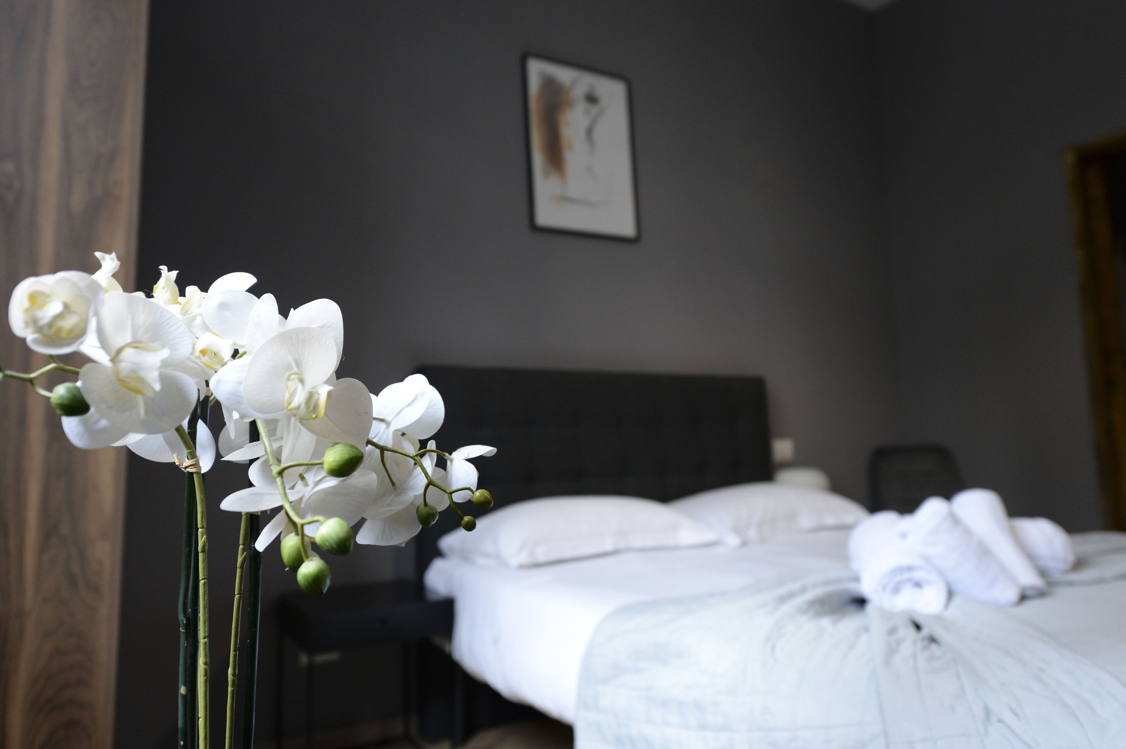 Camera d'albergo con un letto, biancheria da letto bianca, un quadro appeso e un vaso con fiori bianchi.