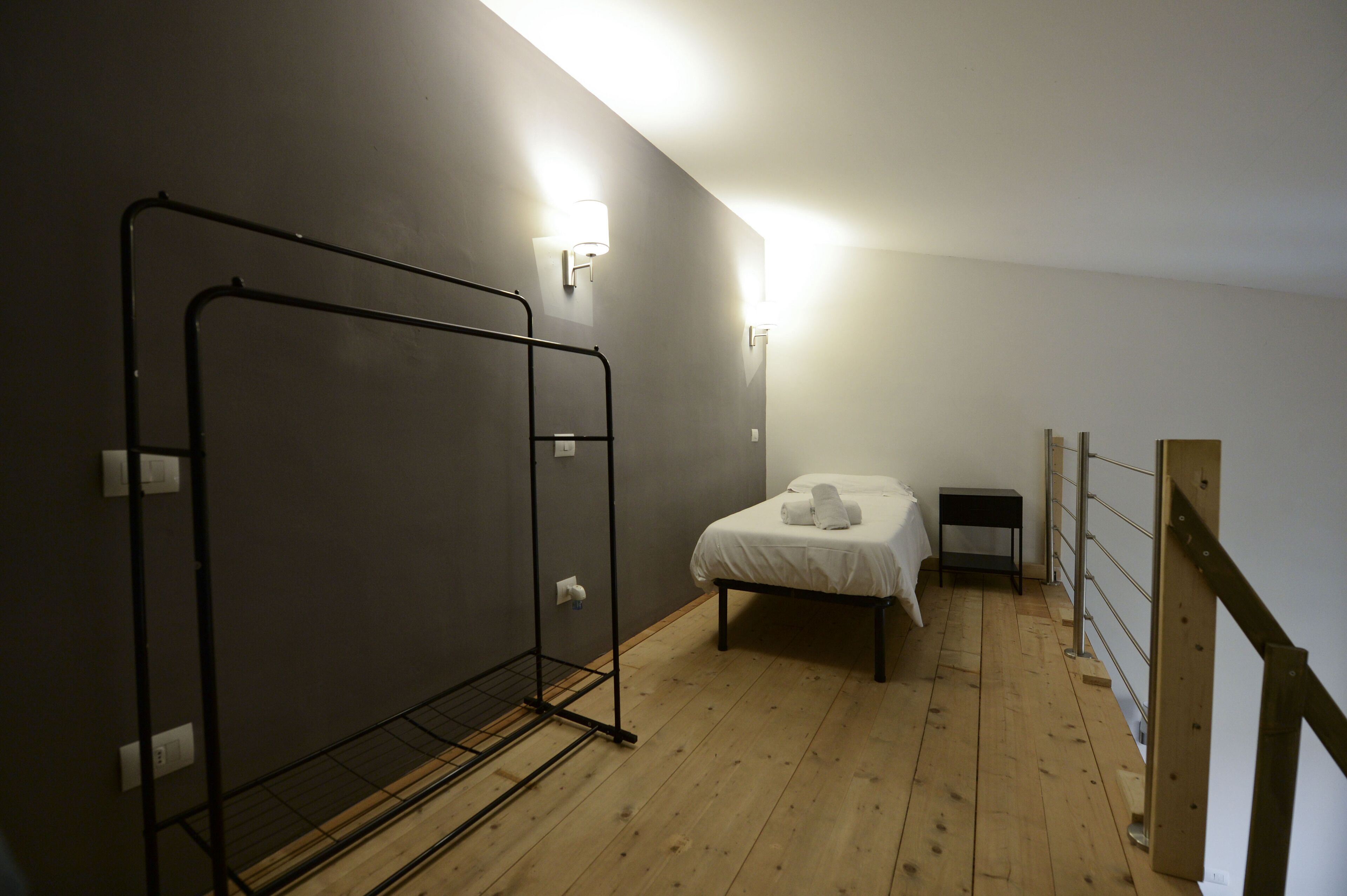 Quarto pequeno e com pouca luz, com uma cama de solteiro, um varal de metal e uma mesinha.