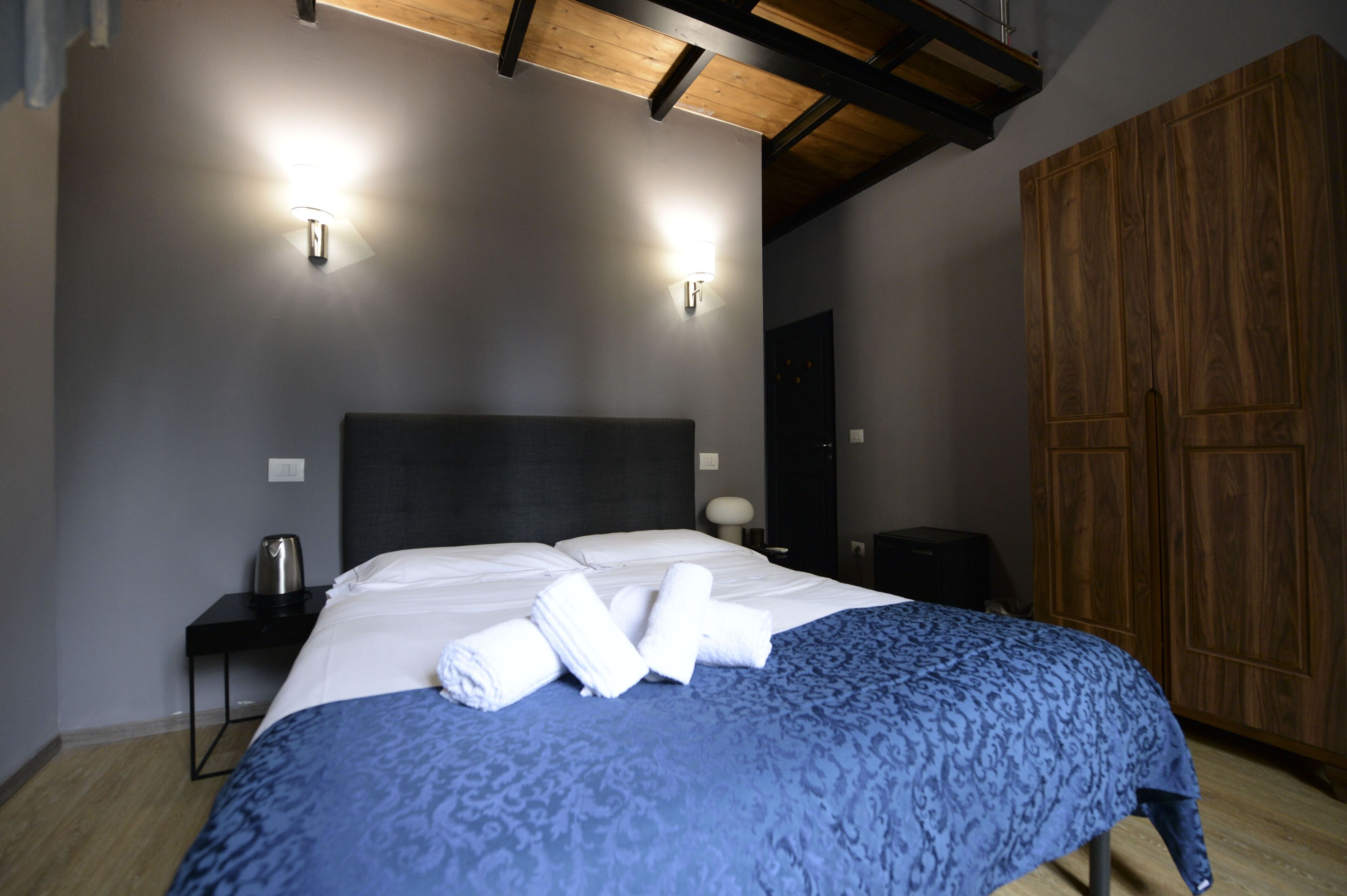 A modern hotel room with a large bed, a wooden wardrobe, and a nightstand with a kettle.