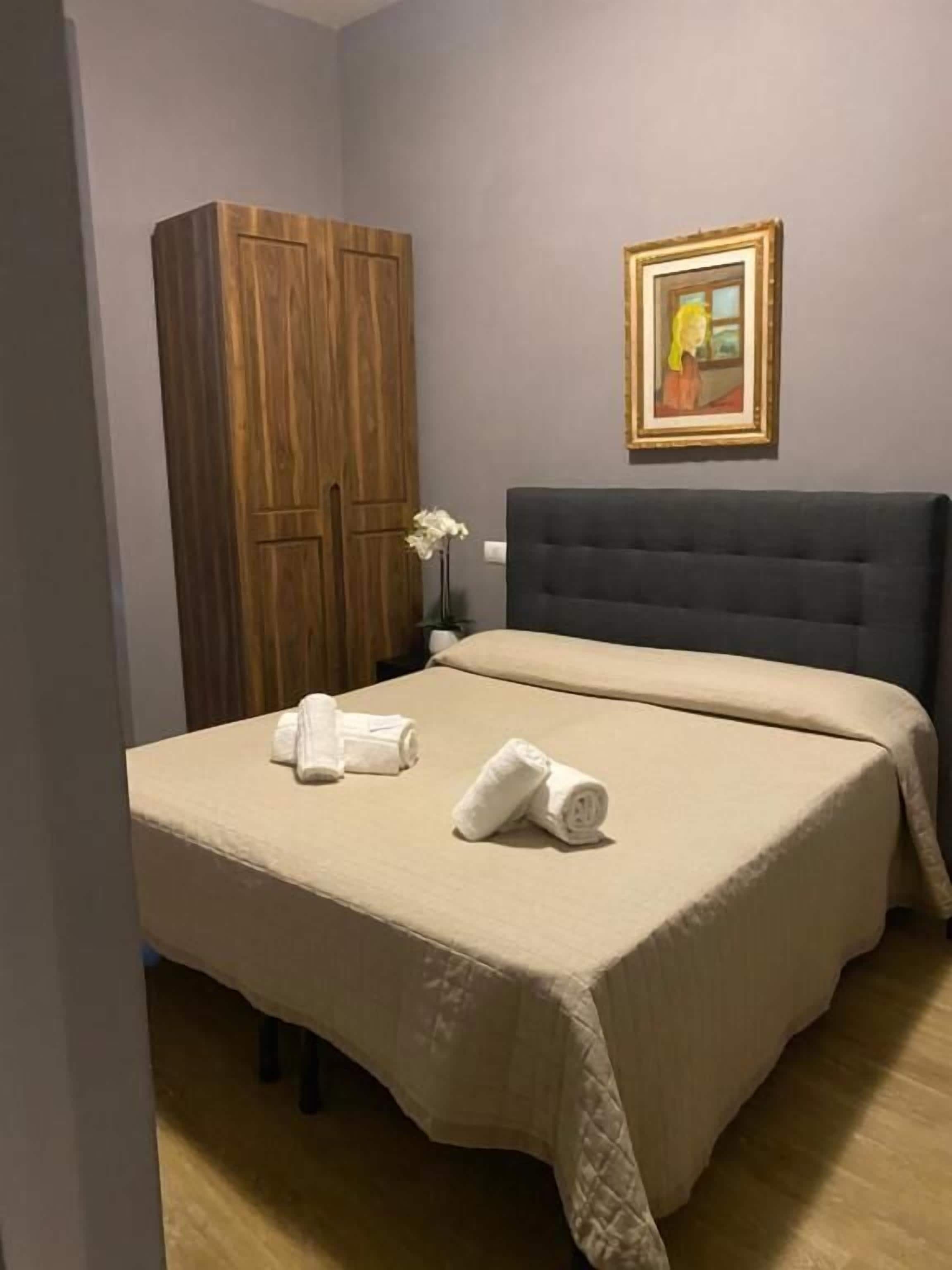 Une chambre à coucher avec un lit, une armoire en bois, un tableau encadré au mur et un vase contenant une fleur blanche.