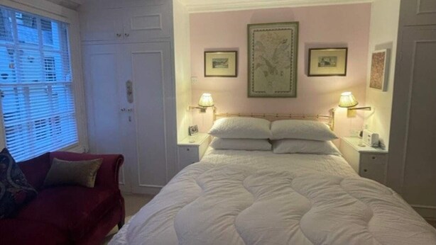 A bedroom with a large bed, two bedside tables with lamps, a red sofa, and a window with blinds. - Expansive 4BD Home- 15 min From Buckingham Palace! (London)