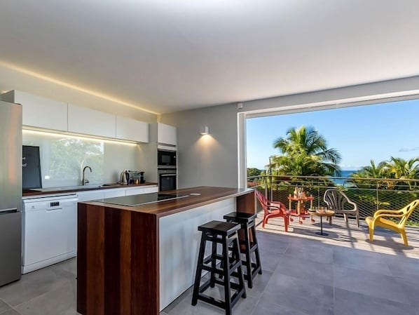 A modern kitchen with a central island and bar stools, a dining area with outdoor seating, and a view of the ocean.