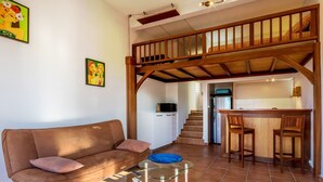 A living room with a brown sofa, a glass coffee table, and a kitchen area with a wooden staircase. - Studio-Romantic-Wet room-Sea view-RDC Avec Terrasse privée (La Trinité)