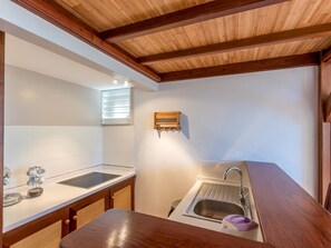 Une cuisine compacte avec un plafond en bois, une fenêtre avec des stores, un évier encastré et un petit plan de travail sur lequel se trouvent une bouilloire et un cuiseur à riz.