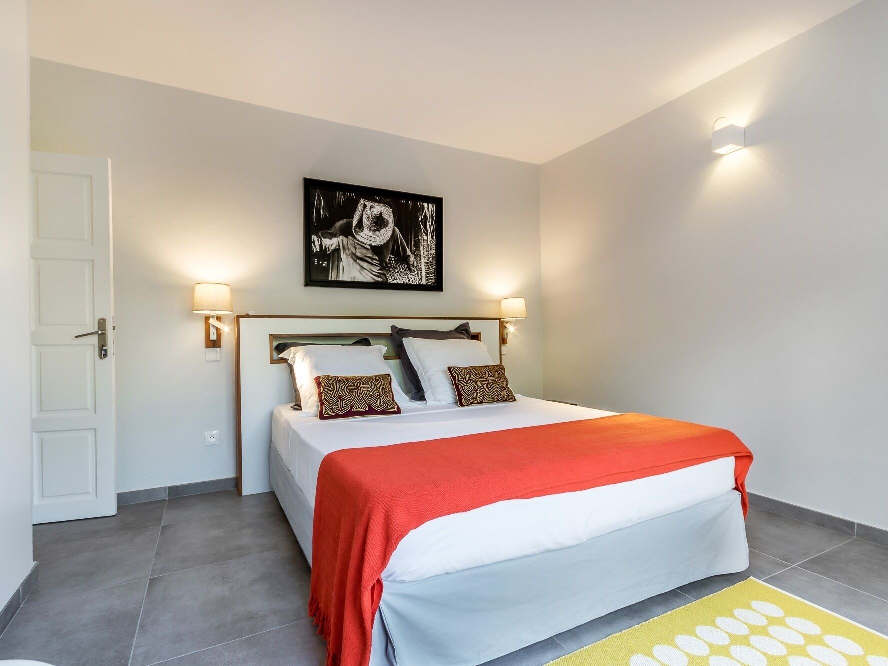 Une chambre d’hôtel avec un lit, une table de chevet, une lampe fixée au mur et un tableau encadré au-dessus du lit.