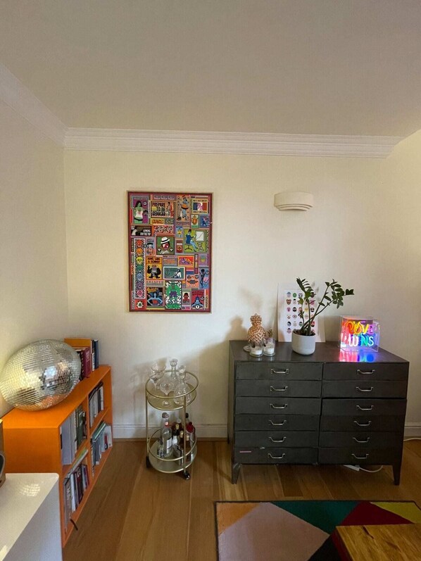 A living room with a bookshelf, a disco ball, a glass cabinet with bottles, a dark dresser, a neon sign, and a colorful poster on the wall.