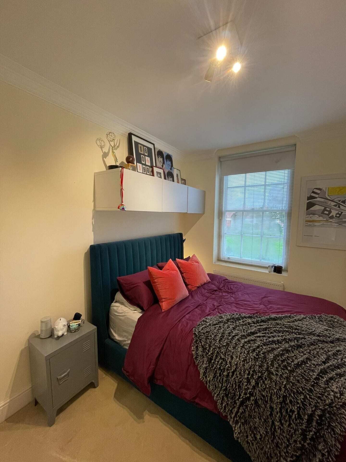 A bedroom with a bed, a nightstand, a window with blinds, and a wall with framed pictures.