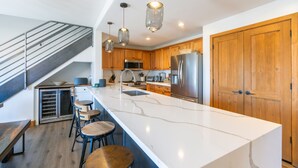 A modern kitchen with a large island, wooden cabinets, stainless steel appliances, and a staircase.