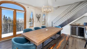 A dining area with a wooden table and blue chairs, a chandelier, a staircase, and a view of snow-capped mountains.