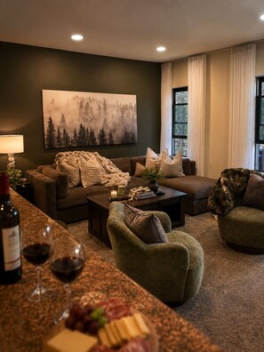 A cozy living room with a dark green wall, a large forest-themed painting, a brown sofa, a green armchair, and a coffee table with books and a candle. - Bear Creek Lodge 303 3 Bedroom Condo by Alpine Lodging Telluride (Telluride)