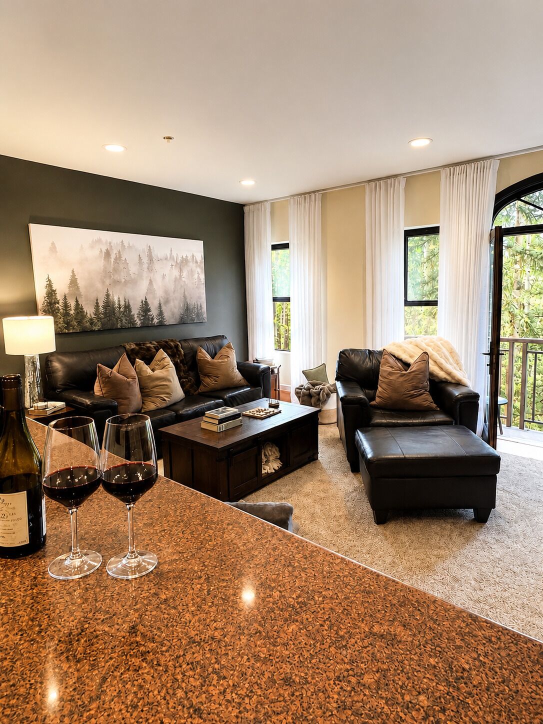 Un salon moderne avec un mur vert foncé, un grand tableau, deux canapés en cuir avec des coussins, une table basse et une bouteille de vin accompagnée de verres.