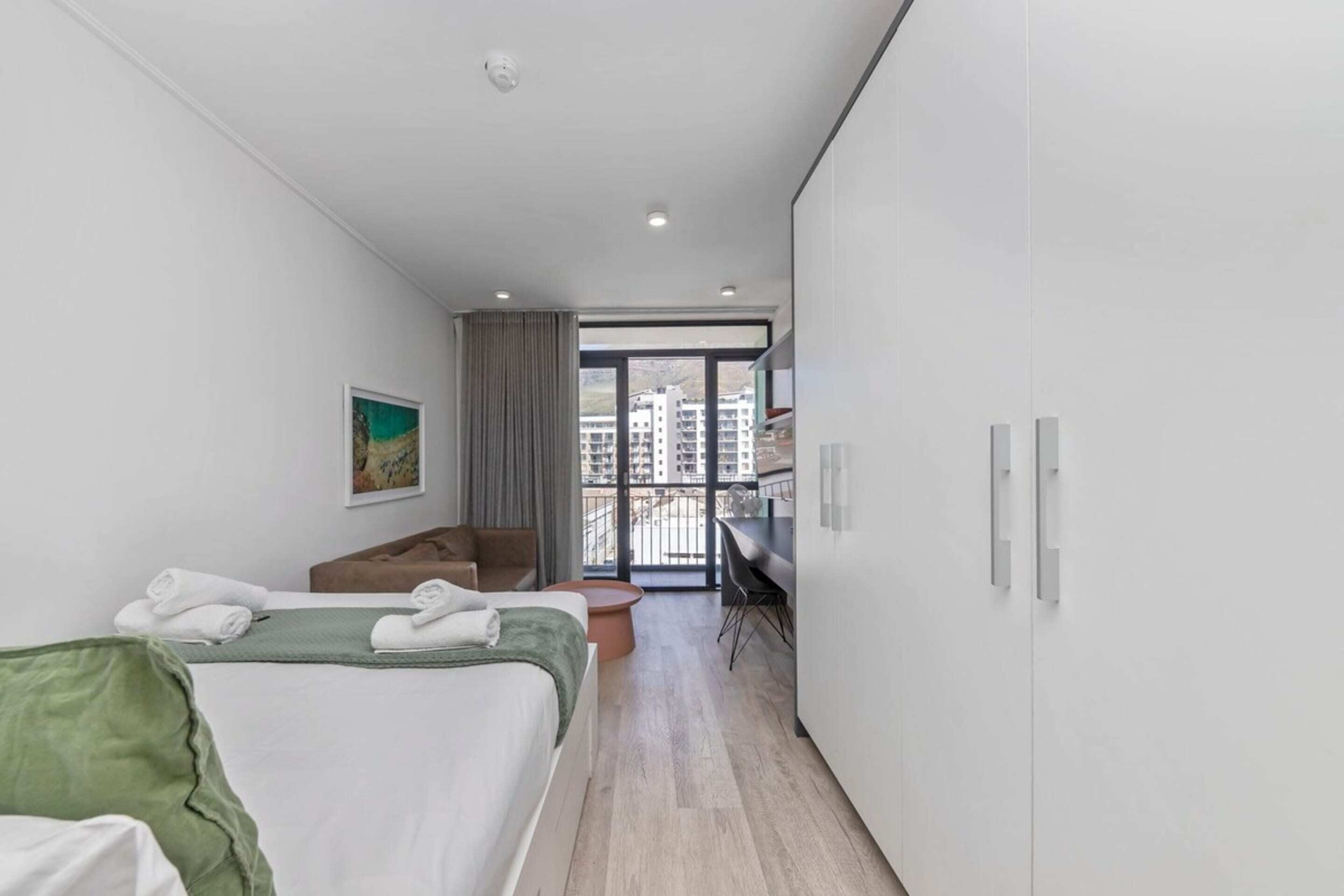 A modern hotel room with a large bed, a desk, and a view of the city through a balcony.