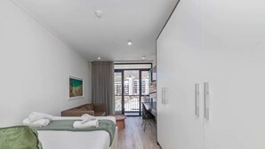 A modern hotel room with a large bed, a desk, and a view of the city through a balcony. - Stunning Studio Apartment With Balcony, Woodstock! (Cape Town)
