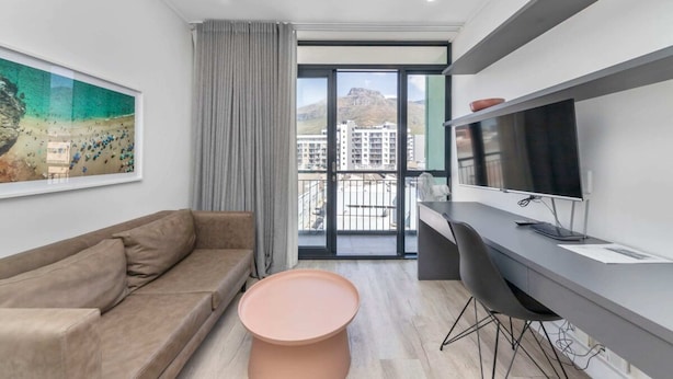A modern living room with a sofa, a round table, a TV, and a balcony with a city view. - Stunning Studio Apartment With Balcony, Woodstock! (Cape Town)