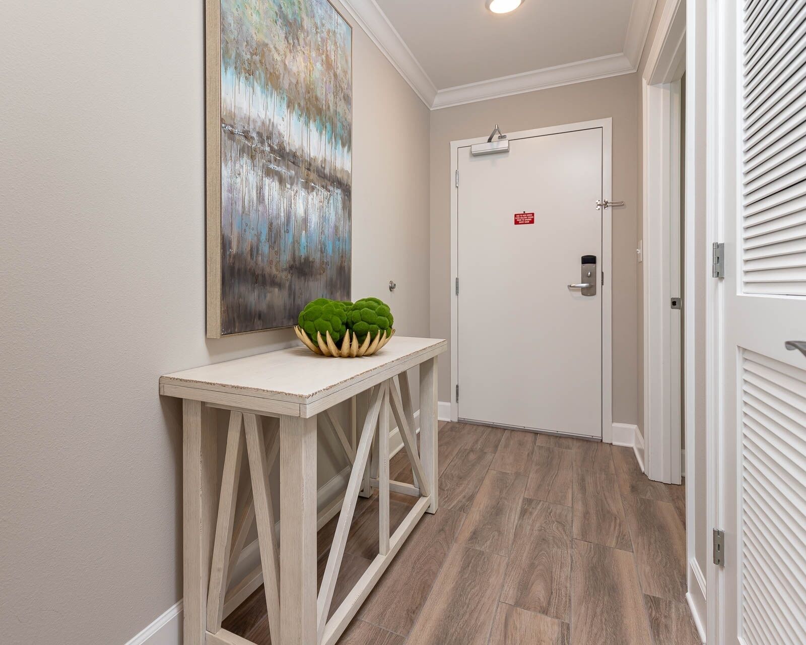 A hallway with a wooden floor, a white door with a red sign, and a painting on the wall.