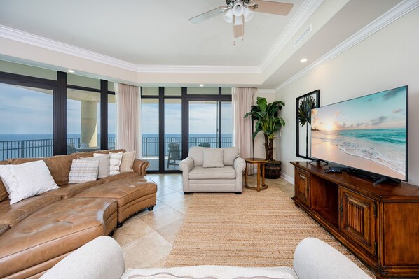 A spacious living room with a large window, a leather sofa, a white armchair, a wooden cabinet, and a flat-screen TV.