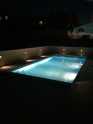 Una piscina rectangular con bordes iluminados, rodeada por una zona de piscina oscura.