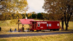 Un wagon de train rouge, typique du Sud des États-Unis, avec un petit bâtiment attenant, un banc et deux chaises, le tout situé dans un espace verdoyant.