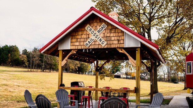 Un kiosque en bois avec un panneau indiquant « PASSAGE PIÉTON », des sièges extérieurs avec des chaises et des tables, et un barbecue.