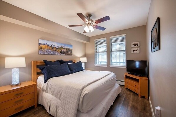 Un dormitorio con cama, ventilador de techo, un cuadro, televisor y mesita de noche.