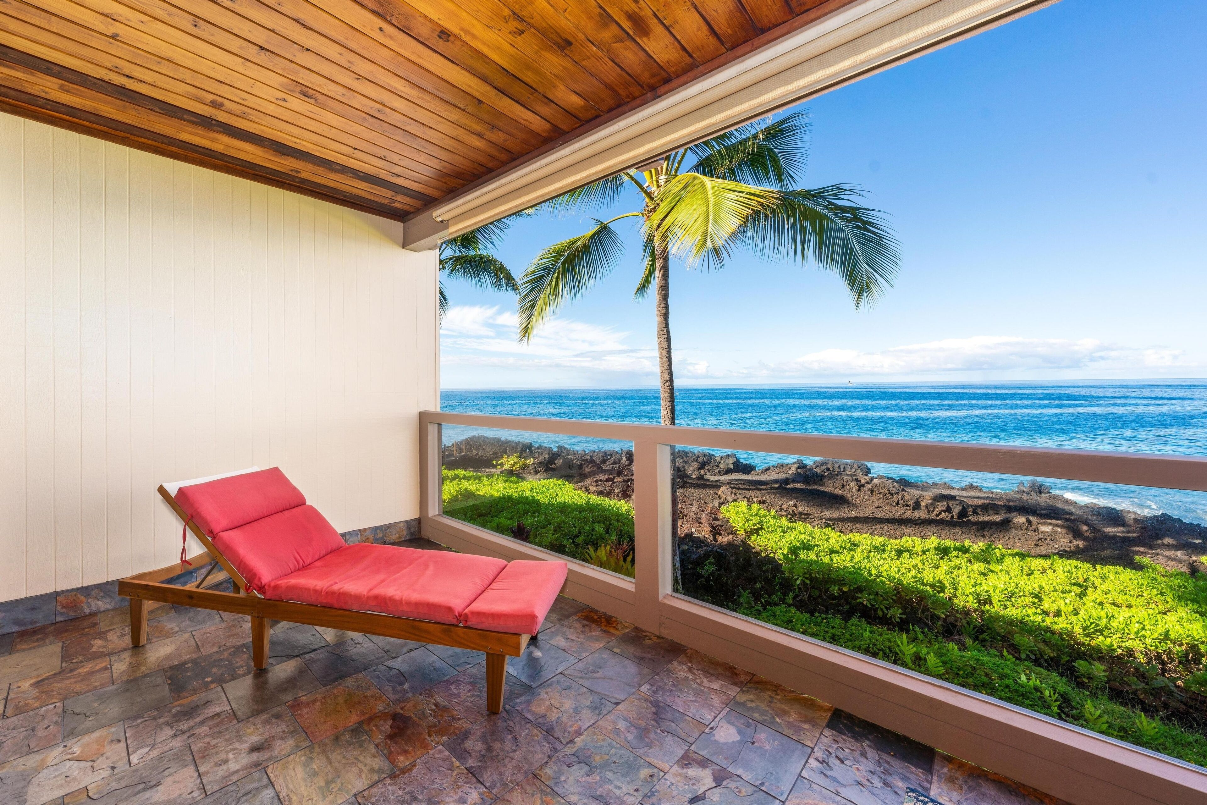 A wooden-framed balcony with a red cushioned chair, overlooking a rocky shoreline with a palm tree and ocean view.