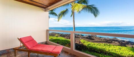 A wooden-framed balcony with a red cushioned chair, overlooking a rocky shoreline with a palm tree and ocean view.