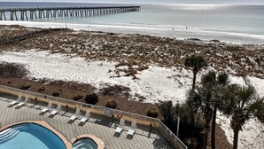 A beach view with a long pier extending into the sea, a sandy beach with sparse vegetation, and a pool area with lounge chairs and a hot tub.