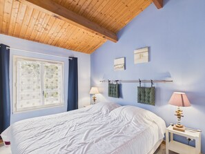 Ein Schlafzimmer mit Holzboden, einem Bett mit weißer Bettwäsche, einem Fenster mit Vorhängen und zwei Wandleuchten.
