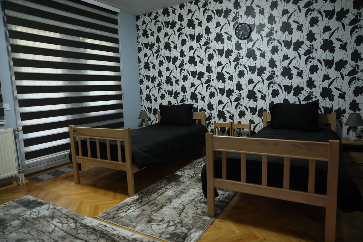 Une pièce avec deux lits en bois, un radiateur, une fenêtre avec des stores et un papier peint à motifs floraux.