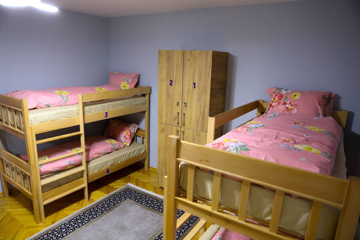 Une chambre avec trois lits superposés, chacun agrémenté de draps roses et de motifs floraux. On y trouve une armoire en bois à deux portes et un petit tapis sur le sol.