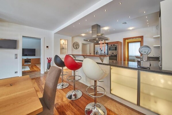 A modern kitchen with bar stools, a dining area with a wooden table, and a built-in oven. - Marys Holiday Penthouse (Saalbach-Hinterglemm)
