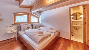 A bedroom with a wooden ceiling, a large window, a bed with white linens, a bedside table, and a bathroom with a wooden door. - Marys Holiday Penthouse (Saalbach-Hinterglemm)