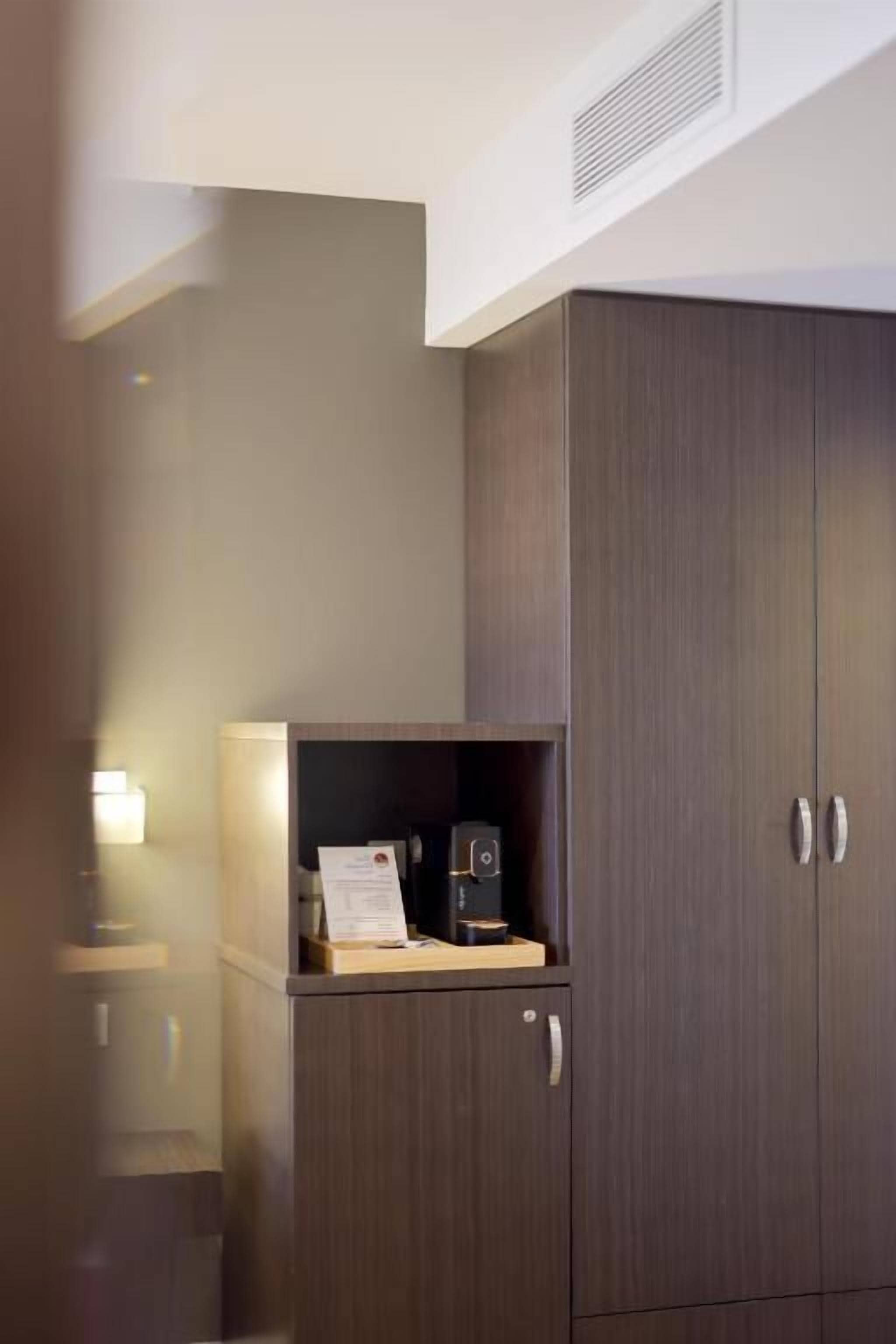 Une chambre d’hôtel avec une armoire en bois foncé, un petit bureau en bois équipé d’une machine à café et un lampadaire fixé au mur.