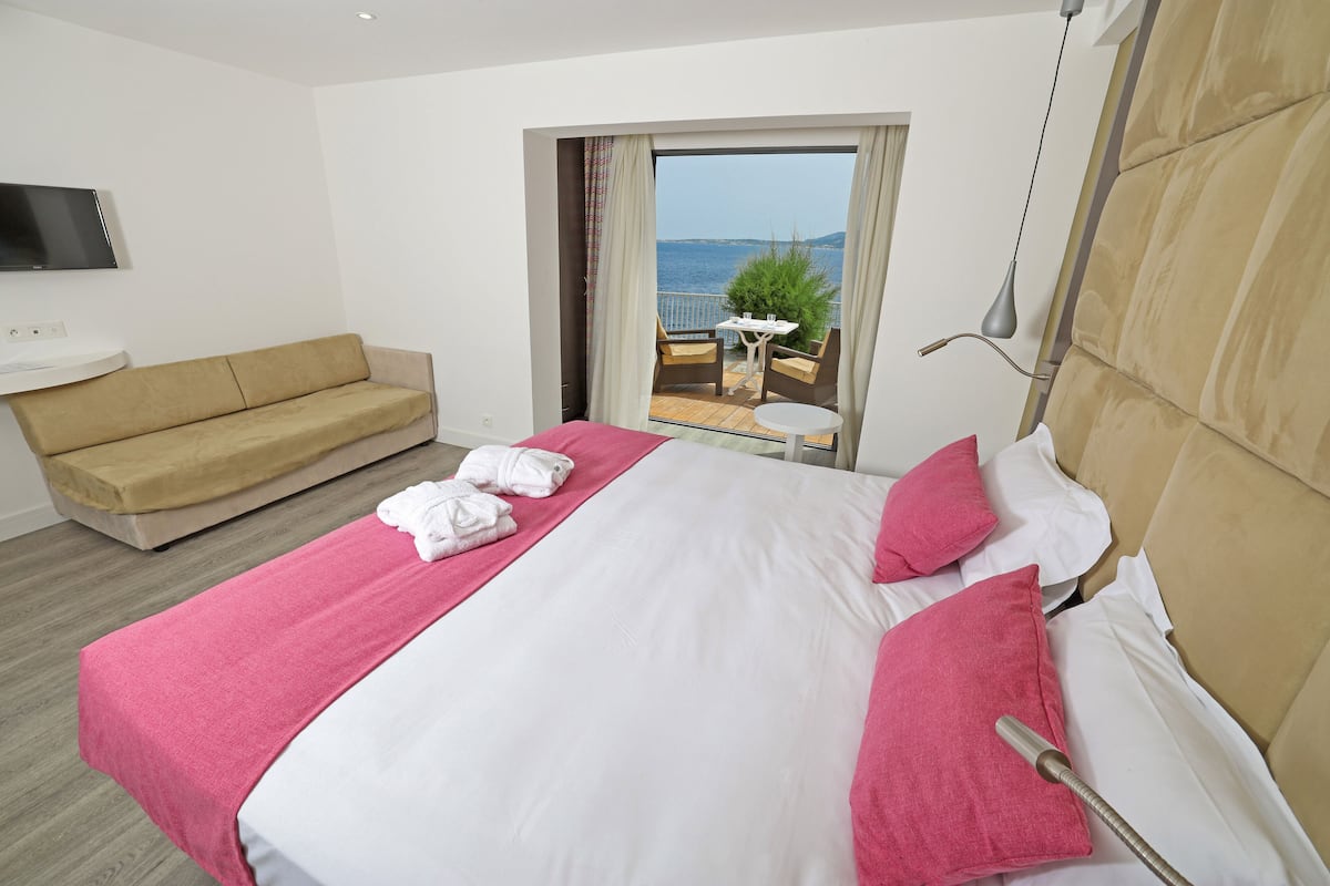 Une chambre d’hôtel avec un lit, un canapé, une télévision, un balcon avec une table et des chaises, et une vue sur la mer.
