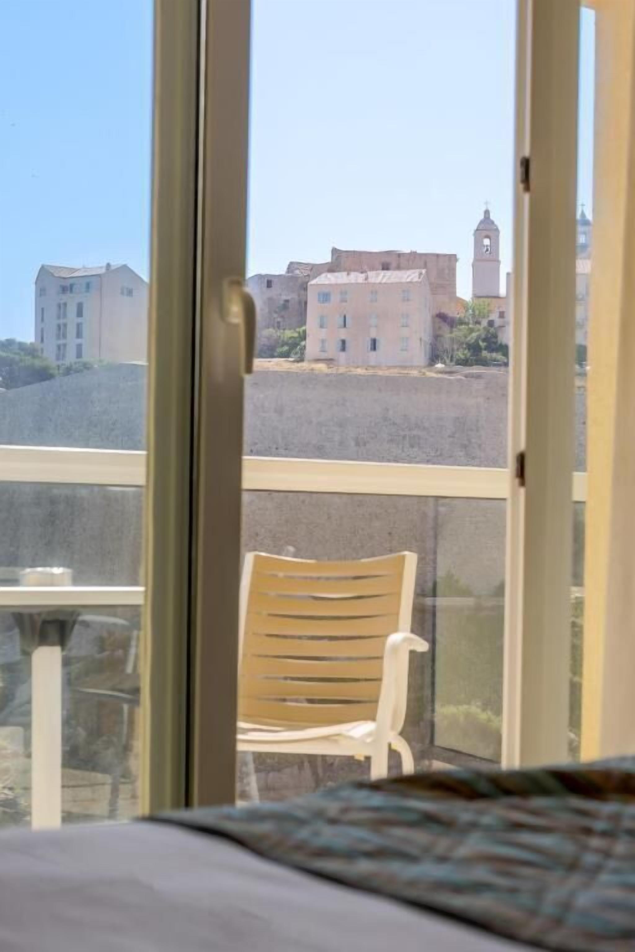 Un balcon avec une chaise en bois, un lit et une vue sur des immeubles et une tour en arrière-plan.